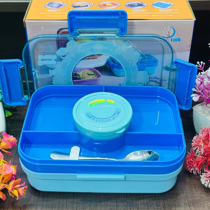 Blue children's lunch box with spoon and lid, featuring a toy inside, on a dark surface with flowers.